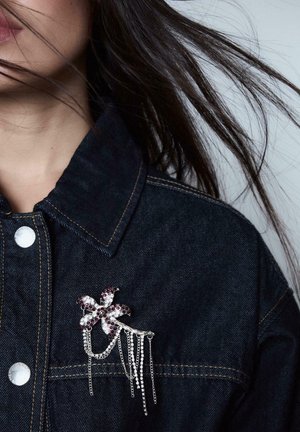 Kvinna som bär en mörk denimjacka med en silver- och lila smyckad blombrosch prydd med hängande kedjor nära kragen.