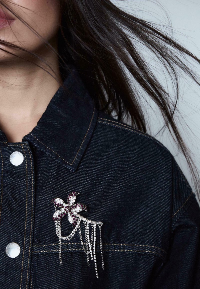 Donna che indossa una giacca di jeans scura con una spilla floreale in argento e viola, decorata con catene pendenti vicino al colletto.