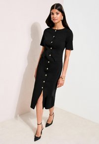 Schwarzes Midikleid mit kurzen Ärmeln, goldenen Knopfdetails und seitlichem Schlitz. Es verfügt über zwei Fronttaschen und ein gerades, strukturiertes Design.