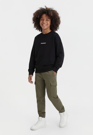 Enfant souriant aux cheveux bouclés portant un sweat-shirt noir DSQUARED2, un pantalon cargo olive et des baskets montantes blanches, debout sur un fond blanc.