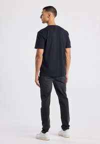 Camiseta de algodón negra con cuello redondo, combinada con jeans oscuros grises. El conjunto se completa con zapatillas blancas, visto desde la parte posterior.