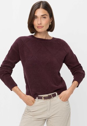 Vrouw met kort bruin haar die gouden oorringen draagt, een donkerpaarse gebreide trui en lichtbeige broek met een bijpassende riem.