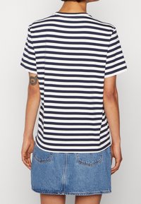 Person som bär en marinblå och vit randig kortärmad skjorta och en blå denimkjol, sett bakifrån med en synlig armtatuering.