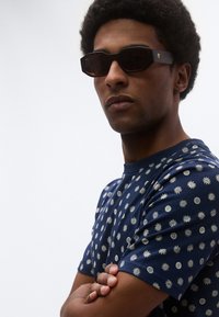 Γυαλιά ηλίου με σκούρους φακούς και εξαγωνικό σχήμα, συνδυασμένα με μπλουζάκι σε σκούρο μπλε χρώμα που έχει ασημένια σχέδια ήλιου.