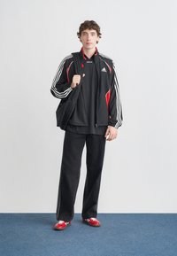 Melns džaka ar sarkanām detaļām un baltām svītrām, melna krekla, plašas melnas bikses un sarkani sporta apavi ar baltām svītrām. Modelis tur melnu somu.