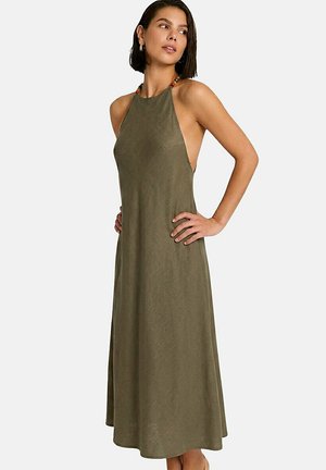 Femme portant une robe sans manches kaki à col halter avec côtés ouverts et encolure perlée, debout les mains sur les hanches, regardant sur le côté.