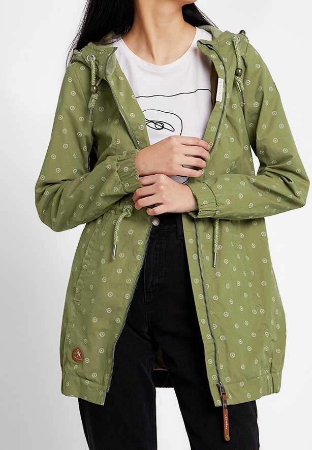 Green, lightweight jacket with a hood, featuring a repeated circular pattern, drawstrings, and a zippered front. Black pants visible.