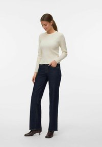 Cremig geripptes Langarmoberteil, kombiniert mit dunkelblauen geraden Jeans. Die Jeans haben sichtbare Nähte und vordere Taschen. Braune Stiefeletten runden das Outfit ab.