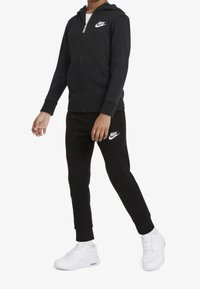 Sudadera negra con cremallera y logo blanco de Nike, combinada con pantalones de chándal negros que presentan un logo blanco de Nike y zapatillas blancas. Textura suave.