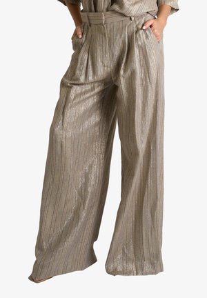 Pantaloni beige a gamba larga con righe verticali e leggero scintillio, mani infilate nelle tasche, abbinati a un top dello stesso tessuto.
