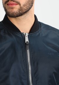 Un homme avec une barbe portant une veste bombardier bleu marine fermée, avec un col en côte noir et un tirette en argent.