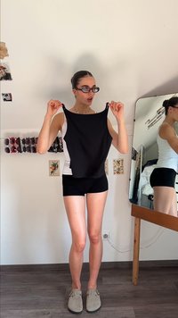 Black sleeveless top displayed by a person, featuring a smooth texture and an elongated cut. Background includes a mirror and wall decor.