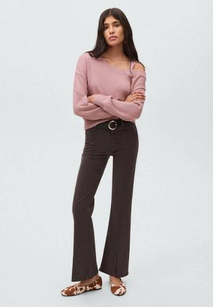 Eine junge Frau steht mit verschränkten Armen da, trägt einen hellrosa, schulterfreien Pullover, dunkelbraune Schlaghosen und gemusterte Ballerinas vor einem grauen Hintergrund.