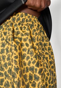 Calções amarelos com um padrão de impressão de leopardo verde, com cintura elástica e tecido texturizado. Camiseta preta parcialmente visível na parte superior.