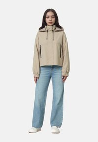 Beige kort jacka med hög krage, dragsko på huvan och två zip-fickor framtill. Kombinerad med ljusblå vida jeans och vita skor.