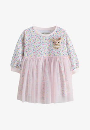Glunderend peutermeisje met kort bruin haar, gekleed in een bloemenjurk en zwarte balletschoenen, staat tegen een achtergrond van roze tule stof.