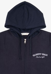 Mørkeblå zip-up hoodie lavet af blødt stof. Har en stor hætte, snore og broderet tekst "VARSITY TEAM" på brystet.