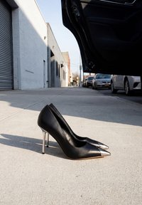 Paire de chaussures à talons hauts noires avec bouts pointus argentés sur un trottoir en ville à côté d'une portière de voiture ouverte et de véhicules garés en arrière-plan.