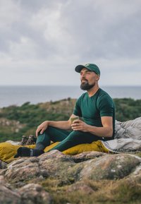 Grünes enges Thermoshirt und Leggings, sitzend auf einem gelben Schlafsack. Hält eine Tasse, trägt eine grüne Mütze, mit einer felsigen Landschaft im Hintergrund.