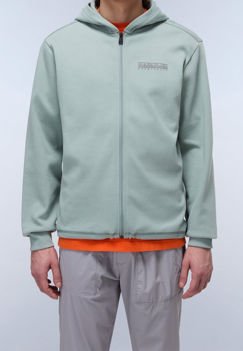 Personne portant un sweat à capuche vert clair zippé avec le logo "NAPAPIJRI", un t-shirt orange en dessous et un pantalon gris clair sur un fond uni.