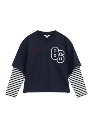 Chemise bleu marine à manches longues, manches à rayures blanches, grand « 86 » blanc et texte rouge « London studio memory » sur la poitrine.