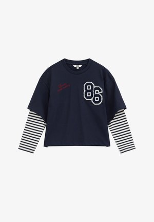 Chemise bleu marine à manches longues, manches à rayures blanches, grand « 86 » blanc et texte rouge « London studio memory » sur la poitrine.