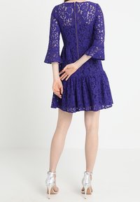 Fialové krajkové šaty s rozšířenými 3/4 rukávy, volánkovým lemem a zapínáním na zip na zadní straně. Kombinováno se stříbrnými sandály na vysokém podpatku.