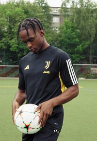 Camiseta negra de la Juventus con acentos amarillos, que presenta rayas horizontales en las mangas. El jugador sostiene un balón de fútbol multicolor en un campo verde.
