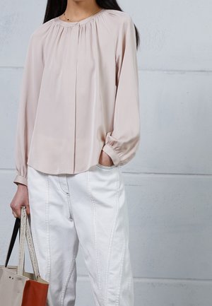Femme portant un chemisier manches longues rose pâle et un pantalon blanc, tenant un sac fourre-tout beige et marron, debout contre un mur gris clair.