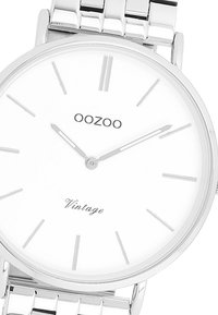 Montre en acier inoxydable avec un bracelet argenté, cadran rond blanc présentant de fins index horaires en argent, et le texte "OOZOO Vintage" en dessous de la position 12 heures.