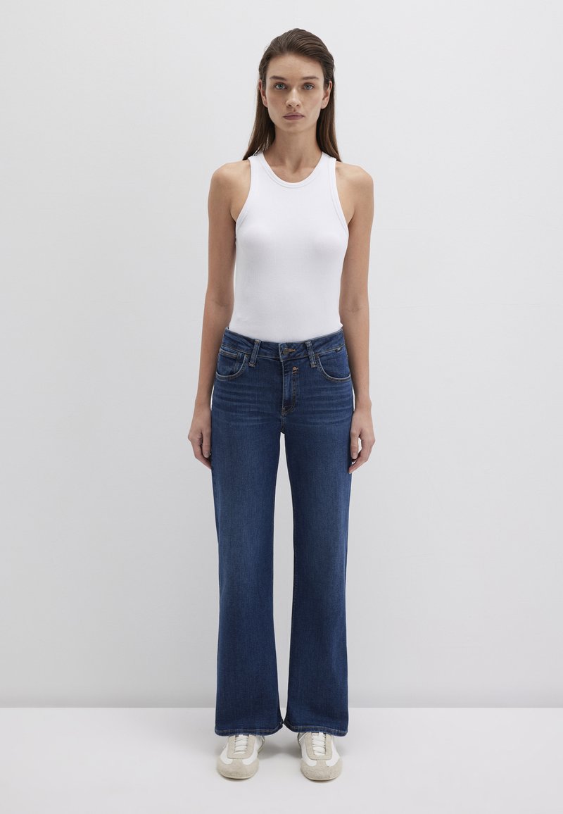 White ribbed tank top paired with medium-wash flared denim jeans. Jeans feature front pockets and a classic five-pocket design. Beige sneakers.