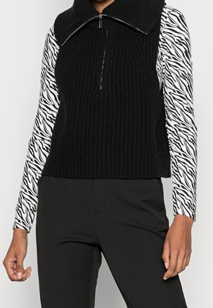 Žena v černém pleteném svetru bez rukávů se zipem přes černobílé zebrované tričko s dlouhým rukávem a černé kalhoty.