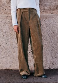 Pantaloni largi din catifea cord cu pliuri în față, culoare verde măsliniu, suprafață texturată și talie înaltă. Asortați cu încălțăminte neagră și un top alb.