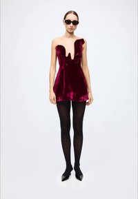 Burgundy velvet mini dress with a deep V-neck, asymmetric shoulder design, and a fitted silhouette. Worn with black tights and pointed shoes.
