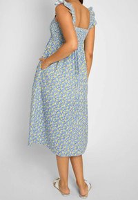 Femme portant une robe midi bleue avec un imprimé floral jaune, des bretelles à volants et des sandales blanches à enfiler, debout devant un fond uni.