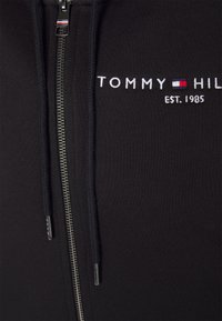 Felpa nera con zip e logo ricamato "TOMMY HILFIGER EST. 1985", zip argento e lacci, realizzata in tessuto morbido con una superficie liscia.