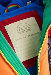 Giacca colorata per bambini con interno in pile blu, tessuto arancione e verde, una toppa rossa con etichetta del nome e zip gialla.