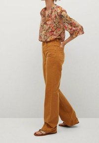 Vrouw in hoge taille, wijde mosterdkleurige broek, bloemenblouse met lange mouwen en tan sandalen, staand met één hand in haar zak.