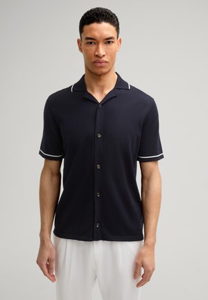 Marineblaues Kurzarmhemd mit Kragen, ausgestattet mit weißen Akzenten und Knopfverschluss. Glattes Material und lockere Passform, getragen mit hellen Hosen.
