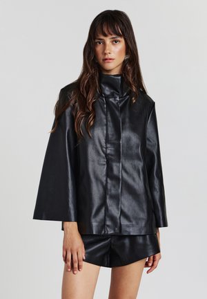 Donna con lunghi capelli scuri che indossa una giacca di pelle nera con maniche larghe e pantaloni corti in pelle nera abbinati, su uno sfondo semplice.