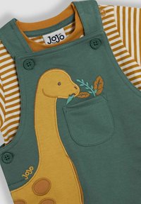 Grüne Latzhose mit gelber Dinosaurierstickerei auf der Vorderseite der Tasche, kombiniert mit einem gestreiften senfgelben und weißen Langarmshirt.