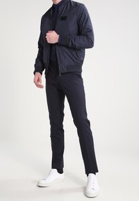 Man in een marineblauwe bomberjack, bijpassende slim-fit broek en witte sneakers, staand tegen een lichte achtergrond.