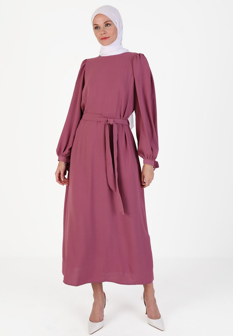 Long, loose-fitting dress in mauve fabric with puffed sleeves and a belted waist. Smooth texture, paired with pointed white heels.