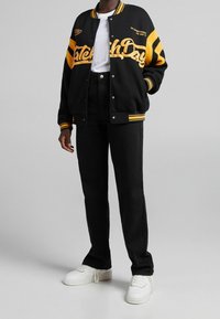 Svart varsityjacka med gula accenter, med texten "Catch Day", bärs över en vit t-shirt och svarta jeans, tillsammans med vita sneakers.