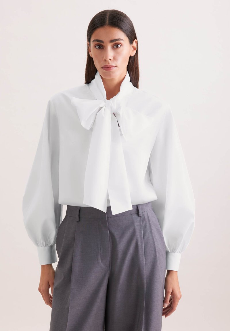 Femme portant un chemisier blanc avec un grand nœud papillon au col et un pantalon gris taille haute à jambes larges, sur un fond uni.