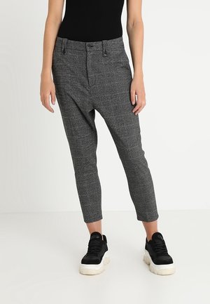 Person iført grå tilspidsede bukser og sorte chunky sneakers stående mod en ensfarvet hvid baggrund.