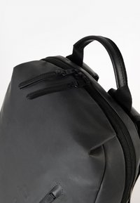 Zaino nero con una finitura liscia simile alla pelle, chiusure a doppia cerniera e manico in tessuto cucito. Presenta accenti di design minimali.