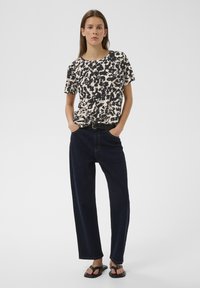 Naine seisab käed taskutes, kandes mustvalget mustrilist lühikese varrukaga särki, tumedasiniseid teksapükse, musta vööd ja musti sandaale.