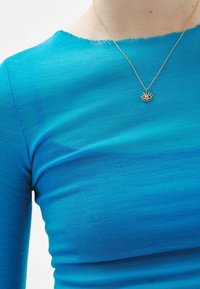 Top azul acanalado con un escote de borde sin terminar, combinado con un collar dorado que presenta un colgante en forma de sol con un diseño de ojo.