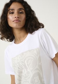 T-shirt en coton blanc avec un grand motif floral beige clair et le texte brodé "FLEURS" au centre. Design à manches courtes et col rond.
